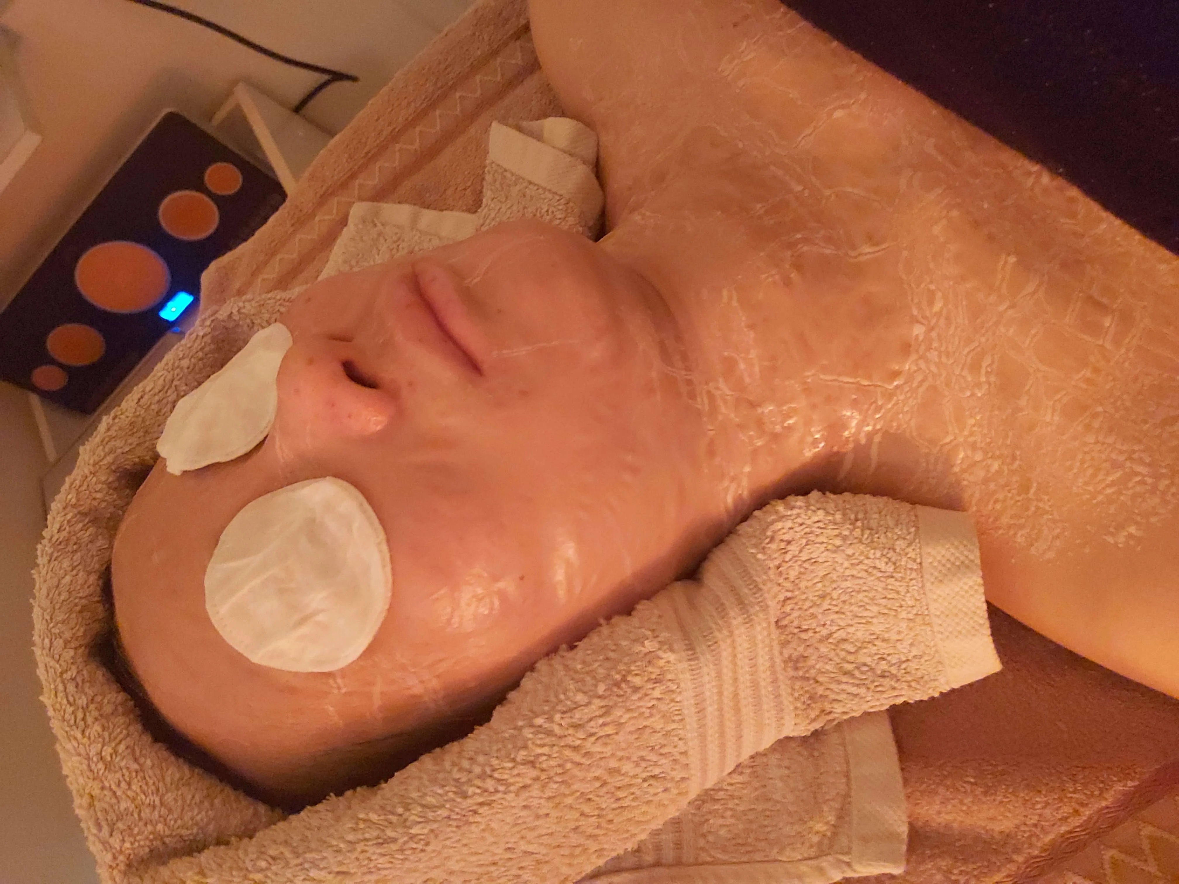 Client reclined under a clear hydrating gel mask with round cotton eye pads and an LED panel glowing at the edge
